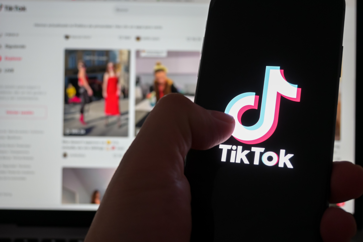 Elde TikTok uygulaması açık telefon, arkada bilgisayar ekranında TikTok video akışı