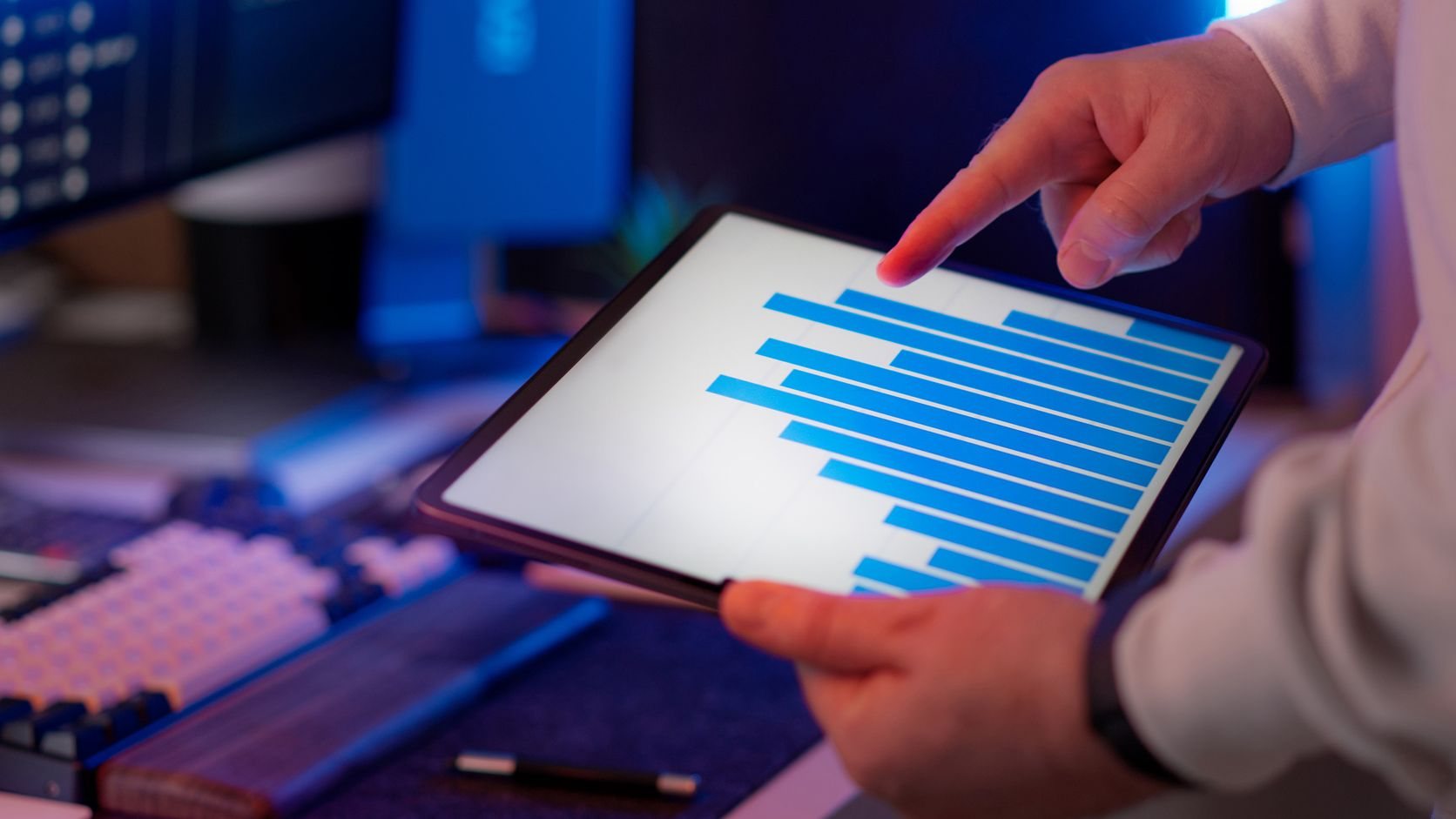 Mavi ışıklı çalışma masasında tablet üzerindeki çubuk grafiği inceleyen el